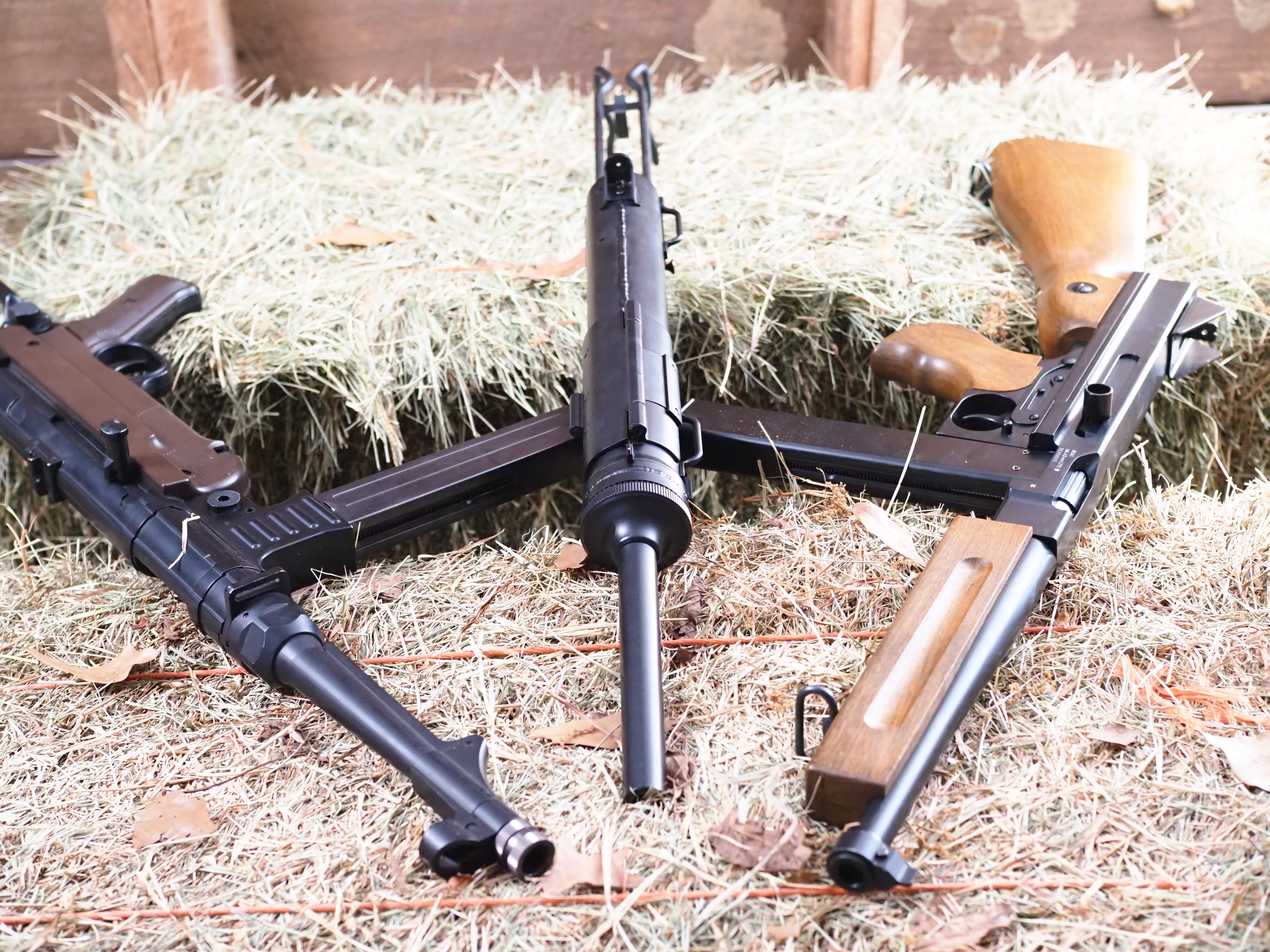 The Legends M3 Grease Gun, MP40, and M1A1 staged on a hay bale